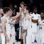 Jugadores del equipo de baloncesto Real Madrid reunidos en círculo en la cancha, levantando sus manos en señal de unión y motivación.
