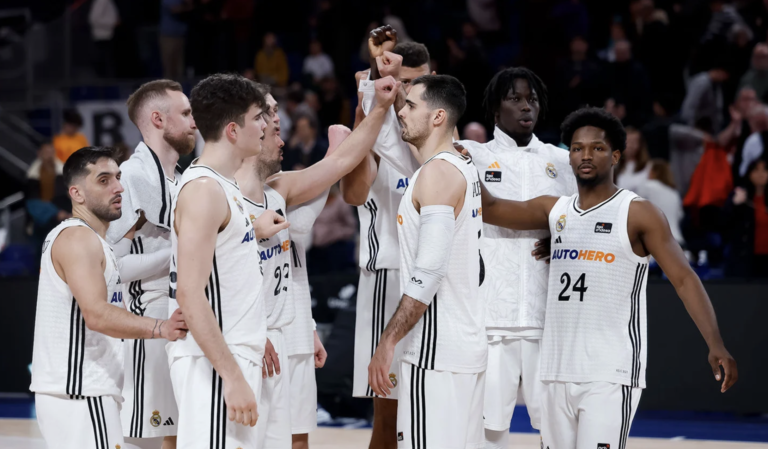 Jugadores del equipo de baloncesto Real Madrid reunidos en círculo en la cancha, levantando sus manos en señal de unión y motivación.