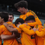 Jugadores juveniles del Real Madrid Catilla celebran un gol abrazándose efusivamente durante un partido contra Algeciras, luciendo el uniforme alternativo de color naranja.