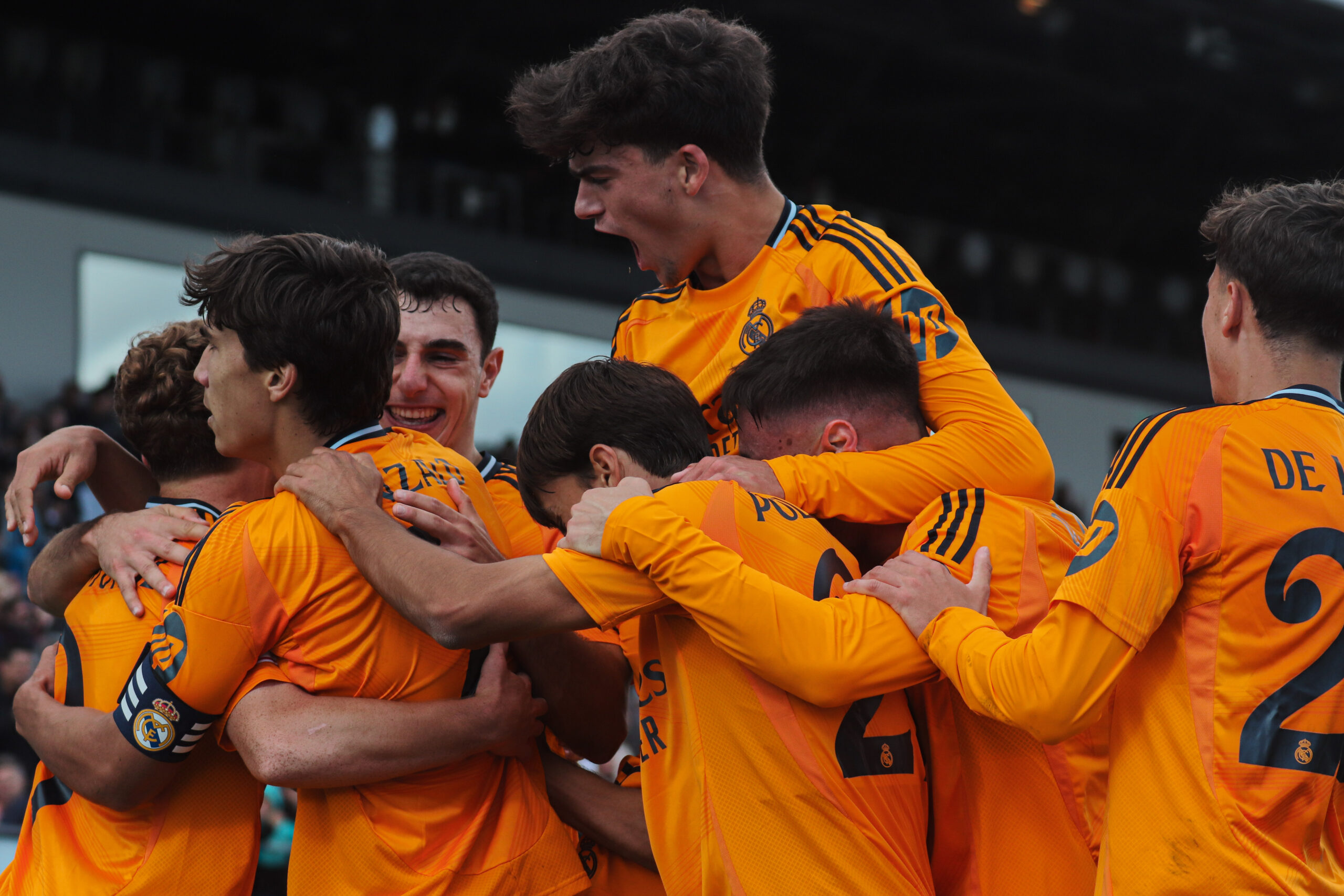 Jugadores juveniles del Real Madrid Catilla celebran un gol abrazándose efusivamente durante un partido contra Algeciras, luciendo el uniforme alternativo de color naranja.